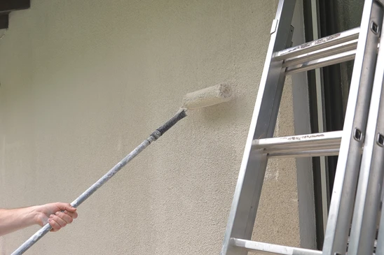 Ext&eacute;rieur, personne appliquant un rev&ecirc;tement sur mur avec rouleau &agrave; manche t&eacute;lescopique pr&egrave;s d'une &eacute;chelle aluminium