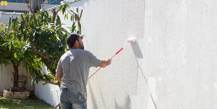 Chantier de construction extérieur avec ouvrier en combinaison bleue appliquant un enduit blanc sur mur béton